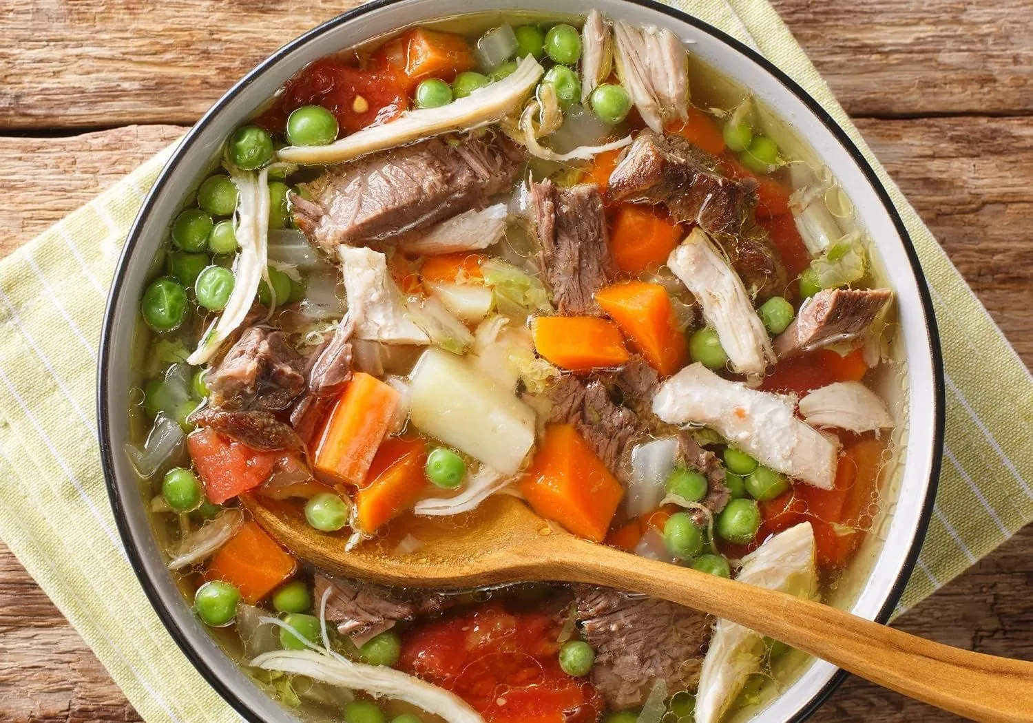 A pot of beef and vegetable soup with peas and carrots, and a wooden spoon in it