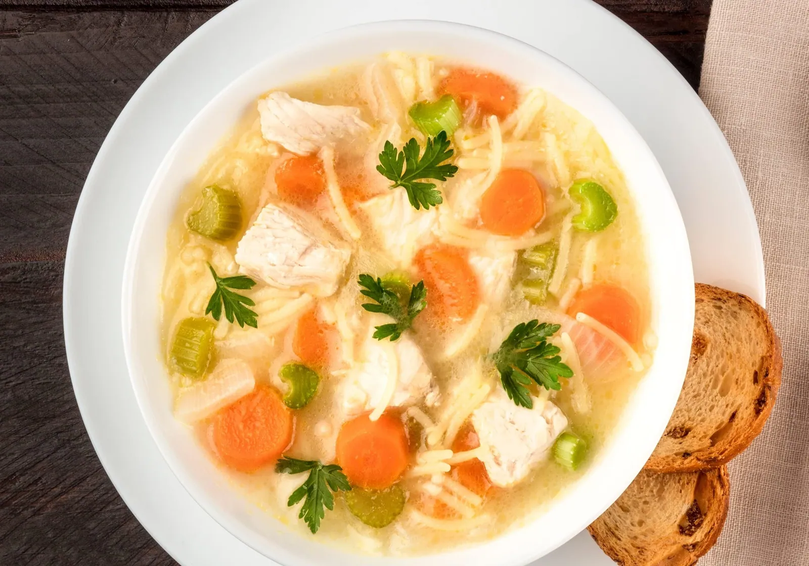 A bowl of chicken noodle soup with carrots, celery and parsley, with toast on the side