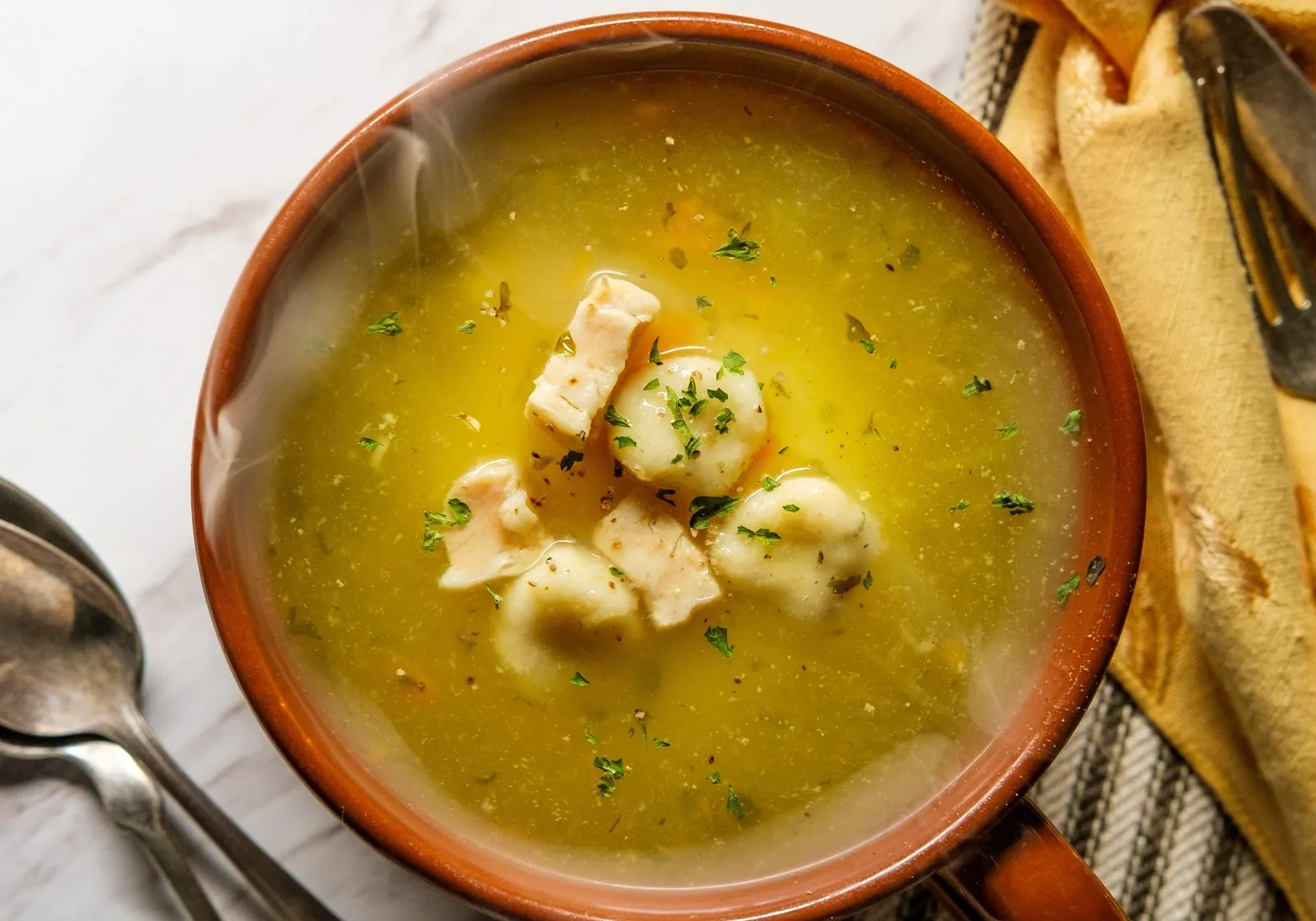 A bowl of chicken and dumplings in broth on a marble table, with spoons and a striped napkin